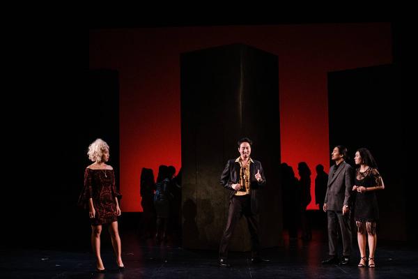 three singers sing stage front, in the background the chorus is silhouetted against a menacing set with a dark red glow.