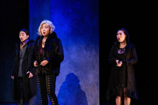 Three performers sing in a triangle formation, against a stark set, washed in lush blue light.