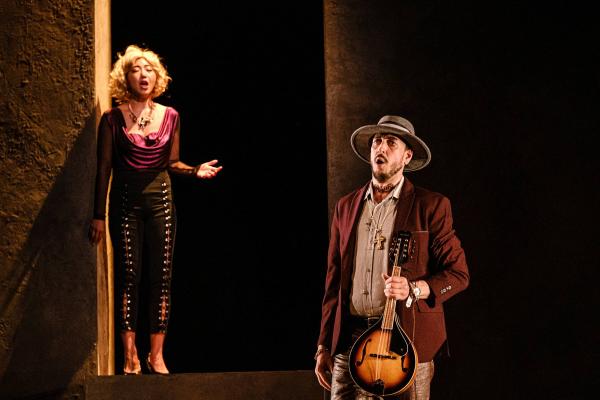 A man in the foreground holds a mandolin, while a woman on an elevated set behind him sings.