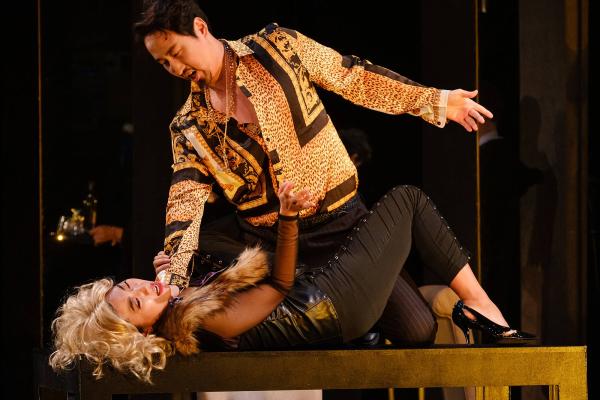A woman lays on top of a table, knees bent, she is clearly in distress, a man stands menacingly above her.