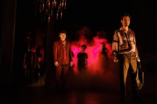 A singer stands defiantly on stage, with fists clenched in anger, while other cast mates look on. In the background several chorus members are silhouetted against and eerie, smoke-fill, blood red background.