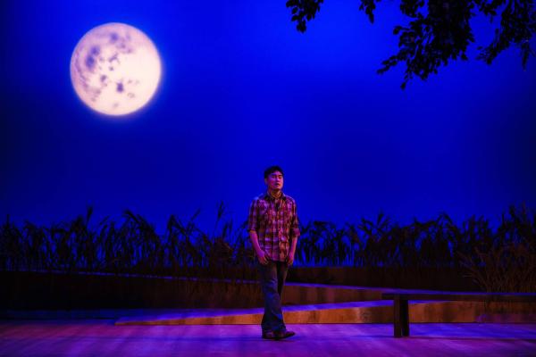 A singer stands alone on a darkly lit stage, with a large full moon behind him.