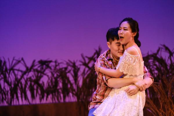 a woman sits on a man's lap as she sings an aria, in front of a wheat field an a lush purple background