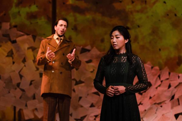 a woman in a black dress stands center stage, with her head turned, listening to man who is standing behind her, singing an aria.