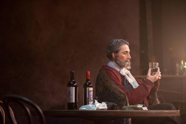 an elderly bearded man sits alone at table staring into a glass of wine. 