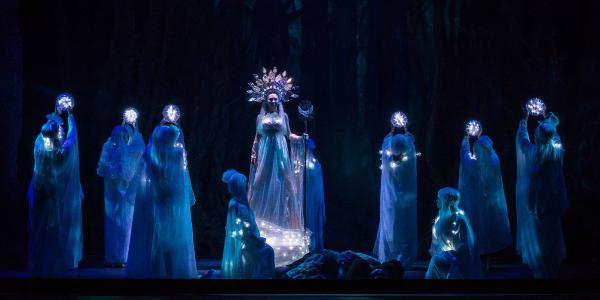 a witch stands on stage in ornate dress surrounded by shadowy figures.