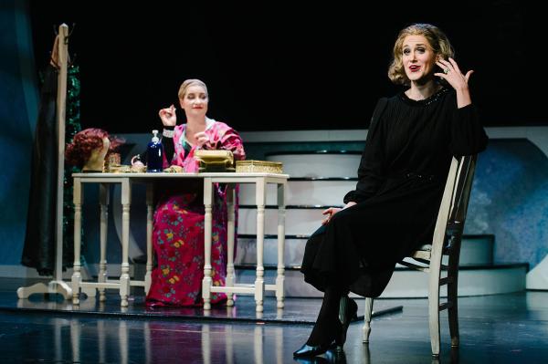 A woman sits sideways on a chair, looking nostalgic, in the background behind her an actress sits at a makeup table, with wig removed, working on her makeup.