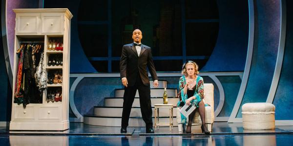 A Man in a tuxedo and a woman in a dressing robe stand near a wardrobe and a night stand.