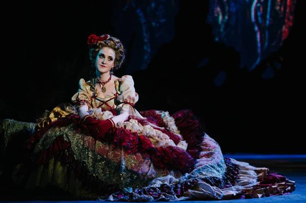 A woman in an ornate Elizabethan dress sits on the ground in a wooded set, looking troubled.