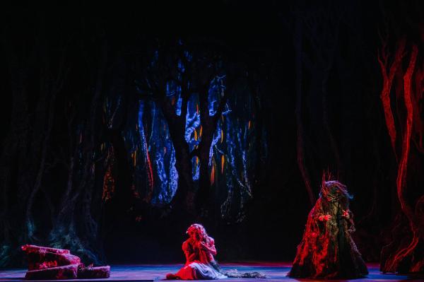 the witch stands hunched over on a wooded set lit by eerie red light while a young girl cowers on her knees, center stage.