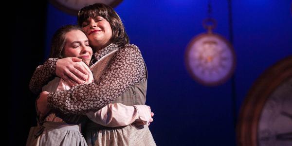 Two women embrace each other with heads close and eyes closed. In the background out of focus, are several large clocks hanging by chains.