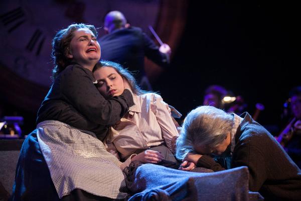 a woman sits on a bed, holding the dead body of young girl to her chest, as a man kneels on the other side of the bed with his face buried in the young girl's lap. 