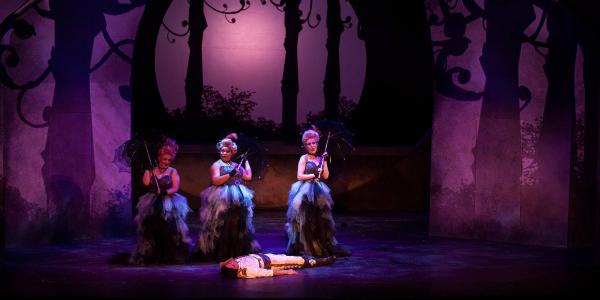 Three women in Elizabethan wigs and dresses stand in an enchanted forest holding parasols, they have stumbled across a man lying unconscious on the ground. 