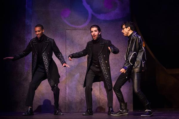 Three men wearing all black jump suits and black leather trench coats plot together in a huddle. 