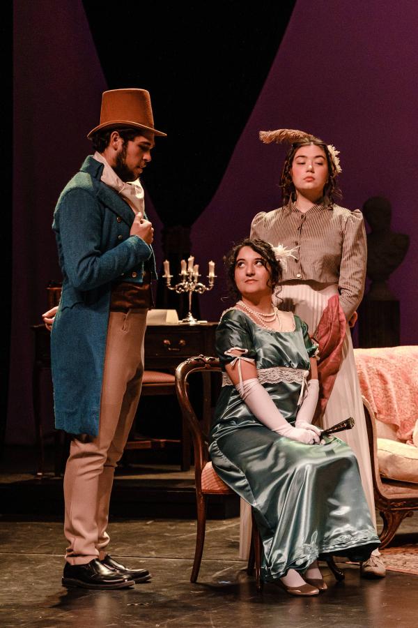 A woman is seated on a chair, flirting innocently with a man standing to her left, dressed in top hat and tails.  An attendant stands behind her, looking unapproving.