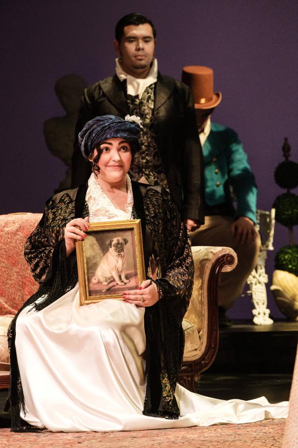 A matronly woman sits tall on a couch proudly displaying a framed portrait of her pug. 