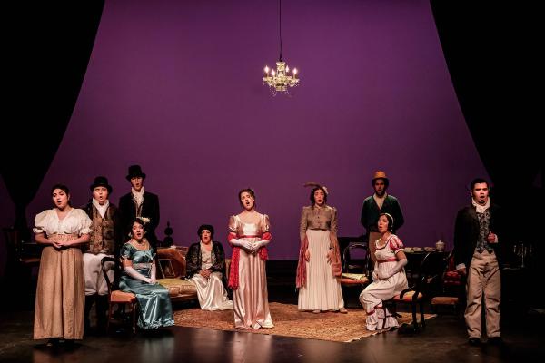 The cast of the opera stand on a minimal set, dressed in formal Elizabethan attire, in a stately formation