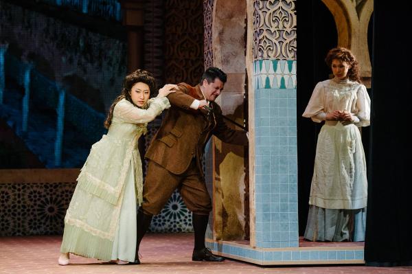 A man tries to break down a door while a woman attempts to hold him back.  On the other side of the door, a woman stands, waiting.