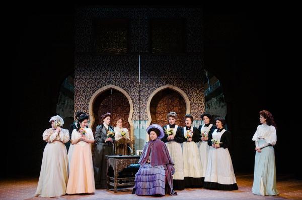 A woman sits somberly on a chair, center stage while house attendants and other women surround her.