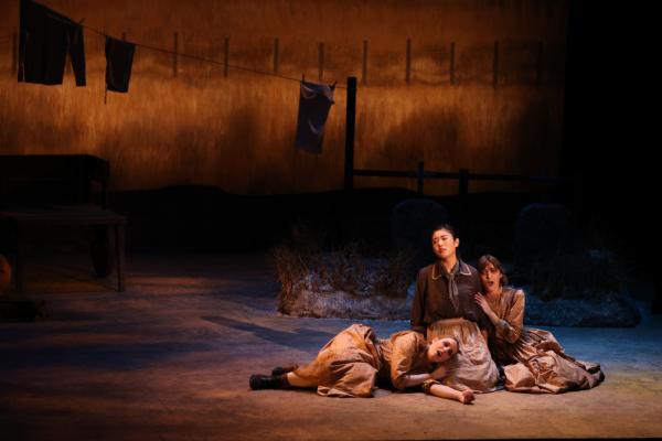 3 women clad in pioneer costumes sit on stage. A central figure appears somber but tall on stage, while another woman leans into her shoulder, and the third lays her head in the strong woman's lap. 