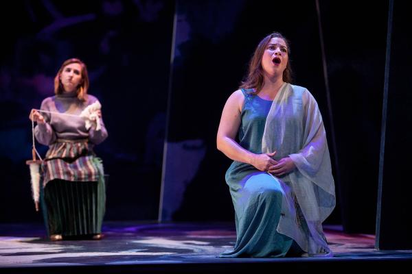 A woman in a flowing Grecian dress kneels on stage, singing in agony. 