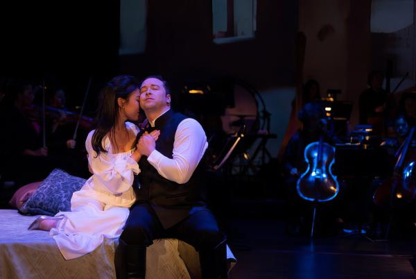 Production still from Shakespeare Gala, a man and woman hold each other closely, seated on a chaise lounge, she buries her head into his shoulder while he looks off into the distance.