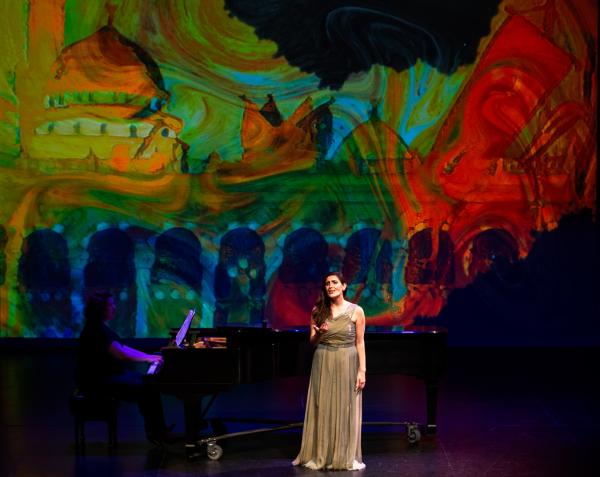 a singer in a formal gown sings on stage, accompanied by a pianist, a bold postmodern background is projected behind them.