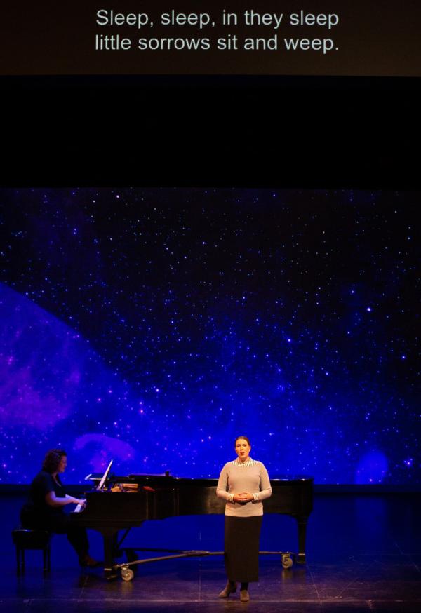 A singer and pianist perform on stage, behind a projection of a clear, starry night. High above them, the supertitles projection reads "Sleep, sleep, in they Sleep, little sorrows sit and weep."