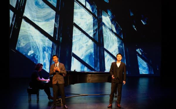 Two singers in dark suits sing on stage, behind them is a projection of a wild electromagnetic storm swirling outside of a giant wall of windows.