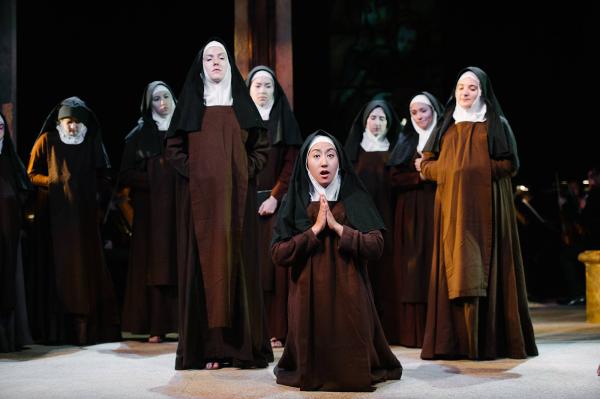 A group of nuns stand center stage. One in front is kneeling in prayer. 