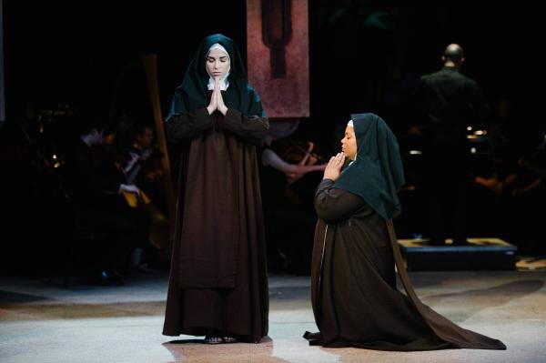 A nun stands prayerfully on stage with her head bowed, while a second nun kneels to her right, facing her, also with hands raised in prayer.