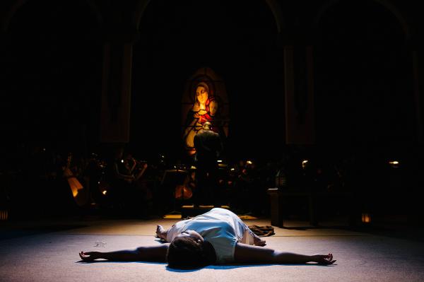 a nun in only undergarments lays splayed out on the stage floor, head toward the audience. She lays unconscious in a pool of light, the faint glow of a stained glass window behind her. The rest of the stage is in total darkness.
