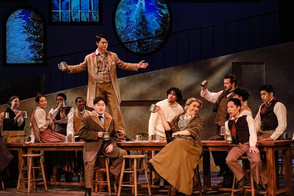 A man dances on a long bar table, while singers clad in victorian peasant clothes lounge in the chairs, drinking beer and enjoying the performance.