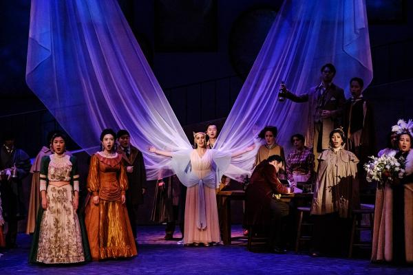 a Woman in and ornate gown and headpiece stand on stage, with huge wing-like spans of cloth extending from her back up into the sky high above the stage. she is surrounded by singes in victorian formal wear.