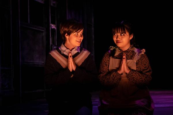 two children kneeled as if to pray, but they look knowingly at each other instead of praying.