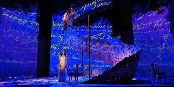 A singer in a white dress stands alone on an expansive set covered in paper, lit with lush blue light.  Cursive writing is projected all over the set. 