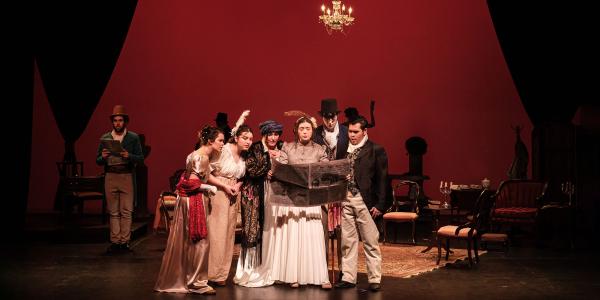 A group of people in formal Elizabethan attire stand center stage, huddled over newspaper that one woman holds up for all.