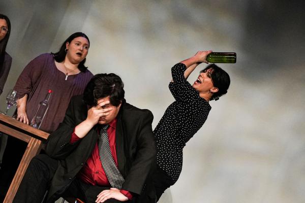 A man sits at a table with his head buried in his hands, while a woman stands behind him, raising a bottle in the air as if about to smash it onto his head.