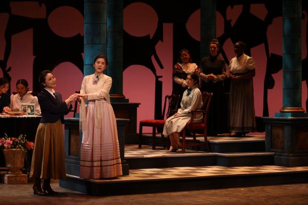 two women sing together, holding hands in front of them, on a darkly lit stage, in the background other performers sit on benches and at tables.