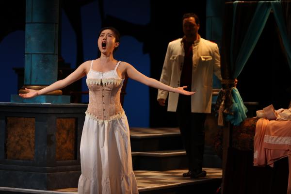 a performer in undergarments sings sorrowfully with arms outstretched, while a man dress in a suit stand ominously behind her.