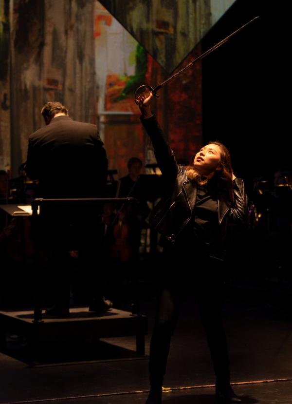 Production Still: A woman clad in black lifts a sword above her head, looking up at the blade in a powerful pose.