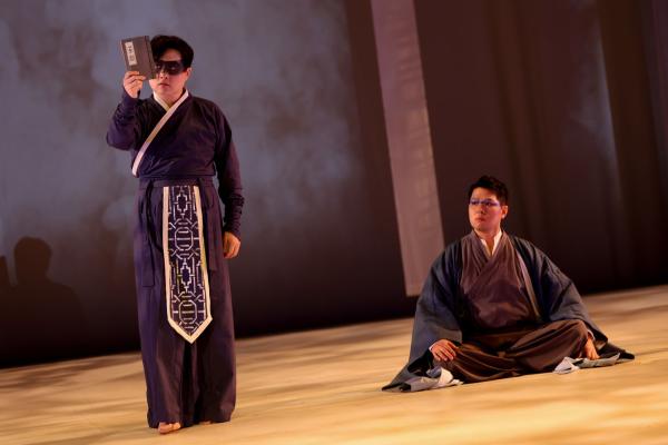 A masked man stands, examining a piece of Parchment while a second man sits cross-legged behind him.