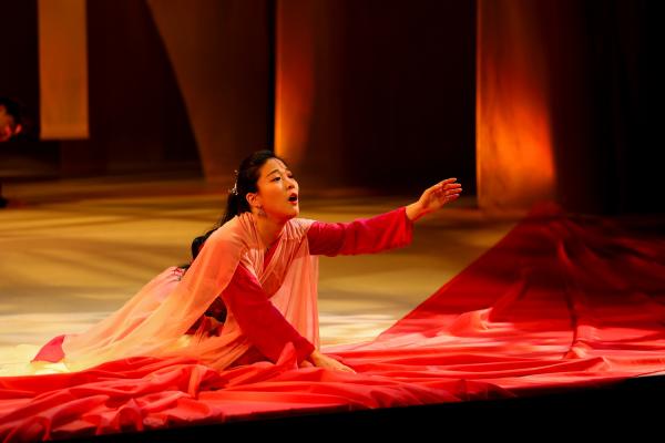 A woman kneels on stage, with an arm outstretched in front of her, surrounded by red fabric, she sings sorrowfully.