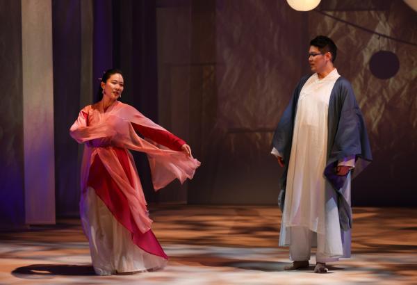 a woman dress in a flowing pink robe dances on stage while a man looks on.