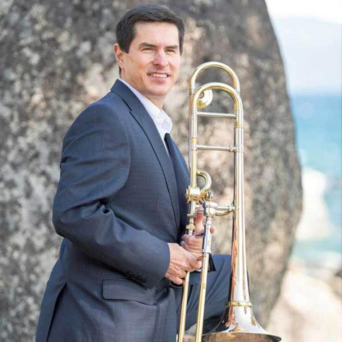 Trombonist David Garcia poses against a cliff with his trombone, the beach an ocean in the distant background.