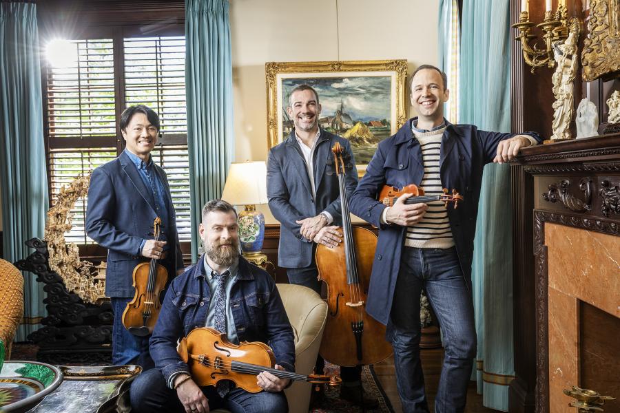 Four mean wearing jeans blue jackets stand in an opulent living room holding their string instruments.