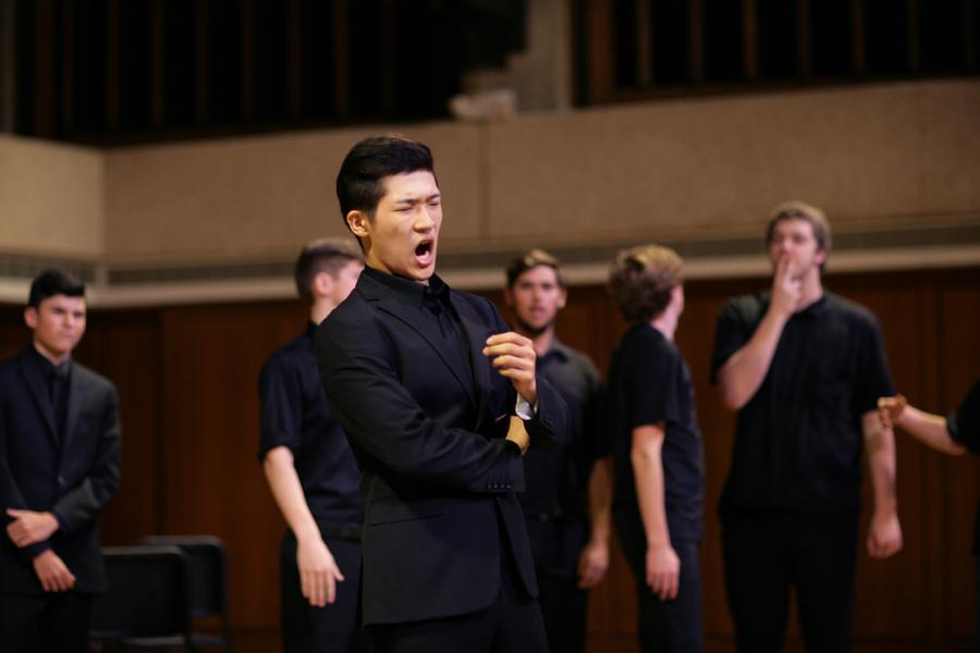An Asian man wearing a black suit sings while crossing his arms. He is surrounded by a group of men on stage.