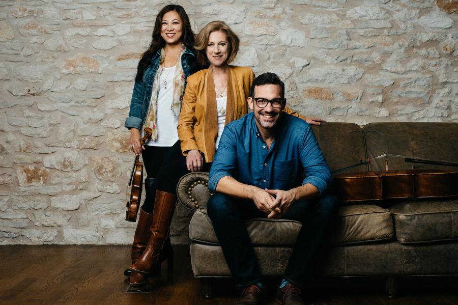 An Asian woman holding a violin stands next to a brown leather couch. To her right, a blonde woman wearing a camel leather jacket leans on the arm of the couch. A man wearing a blue shirt and glasses sits on the couch and a cello lays on the couch beside him.