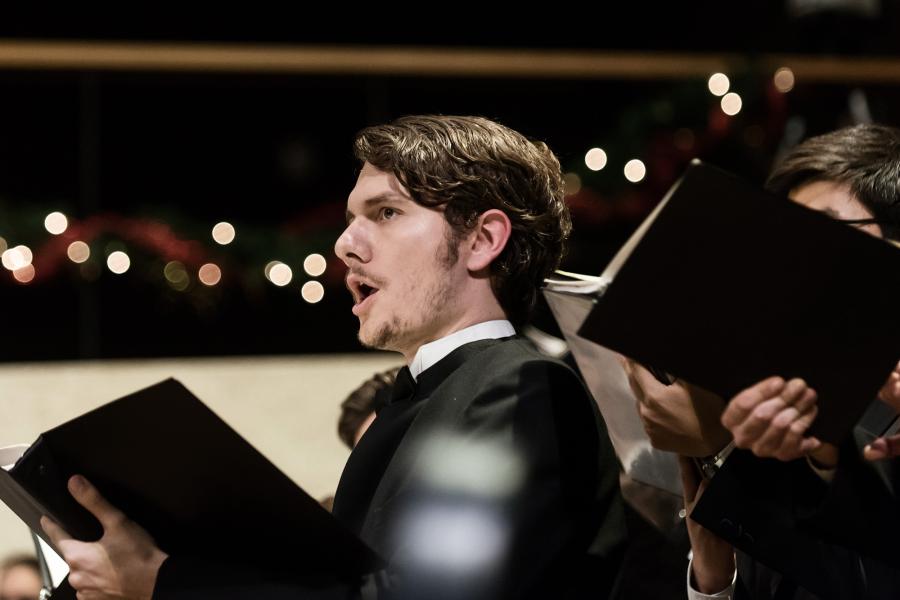 A white man in a black suit sings while gazing forward and holding a folder. 