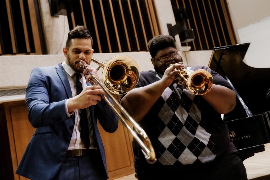 A man wearing a blue suit plays a trombone. To his right, a man wearing a black, white and gray checkered shirt plays a trumpet.